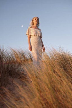 Zarif bir kadın rüzgarlı otların arasında duruyor, ılık günbatımı ışığında, engin, berrak bir gökyüzünün altında, sakin ve güzel bir duyguyu yakalıyor..