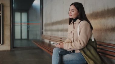 Düşünceli genç bir kadın istasyonda bir bankta tek başına oturuyor, akıllı telefonuyla meşgul oluyor, muhtemelen mesajları okuyor ya da araba bekliyor..