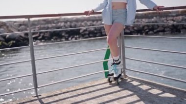 Yazın deniz kenarında paten kaymaktan hoşlanan genç bir kadın, kot şort ve günlük yaz kıyafetleri giyiyor..