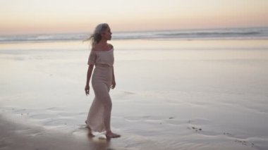 Olgun bir kadın, gün batımında huzur ve özgürlüğü kucaklayarak sahilde huzurlu bir yürüyüşün tadını çıkarır..