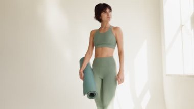 40 'lı yaşlarda güzel esmer bir kadın yoga minderi tutuyor, antrenmana hazır. Güneşli bir stüdyoda yoga kıyafetleri içinde, sağlığı ve sağlığı somutlaştırıyor..