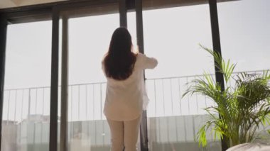 Yeni bir günün ve yeni bir başlangıcın göstergesi olarak içeri temiz hava girmesi için pencereyi açan bir kadının arka görüntüsü..