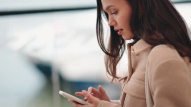Genç bir kadın feribot terminalinde büyük bir pencerenin önünde modern seyahat ve bağlantıyı somutlaştıran akıllı telefonunu kullanıyor..