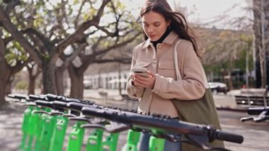 Açık havada akıllı telefonunu kullanarak QR kodunu tarayan ve şehir ortamında elektrikli scooter kiralayan genç bir kadın..