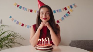 Kapalı gözlü, şenlikli şapkalı bir kadın doğum günü pastasının üzerindeki mumları söndürmek üzere. Renkli kiraz çiçekleriyle çevrili..