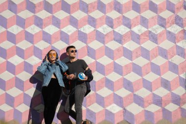 Bir çift, parlak güneş ışığı altında renkli bir geometrik duvara yaslanarak, neşe ve rahatlama yayarak müziği seviyor..