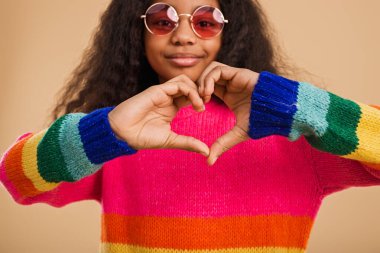 Kıvırcık saçlı bir kızın renkli bir gökkuşağı süveteri ve pembe güneş gözlüğü takıp kendini pozitiflik ve ifade ederken elleriyle kalp hareketi yaptığı yakın plan bir görüntü.