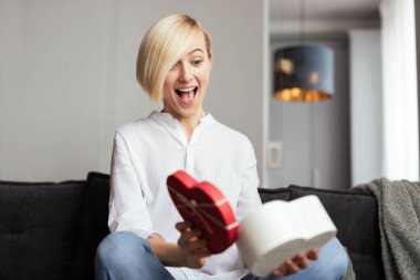 Genç bir kadın mutluluğu ve sürprizi ifade eden kalp şeklinde bir hediye kutusu açıyor. Mekan rahat ve modern. Sevgililer Günü gibi özel bir gün ya da düşünceli bir kutlama gibi.