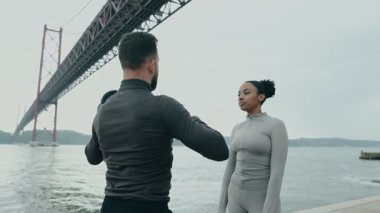 Sportif bir adam, deniz kenarındaki spor salonunda arkadaşlarıyla sohbet ederken dambıllarla omuz egzersizi yapıyor..