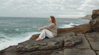 Düşünceli bir kadın denizin kenarındaki kayalık bir uçurumda oturur, uzaklara bakar, çevresindeki okyanus esintisiyle hayatı yansıtır..