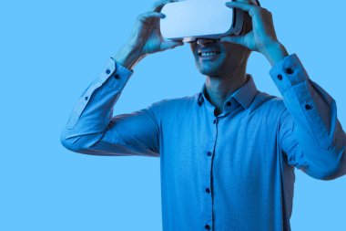 Mavi gömlekli bir iş adamı sanal gerçeklikle etkileşime geçiyor. VR gözlükleri takıyor, gülümsüyor ve nişanlı. Canlı mavi arkaplan, bu resimde aktarılan teknolojik ve gelecekçi kavramı geliştirir.