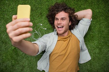 Cheerful young man enjoys a moment of laughter while lounging on vibrant green grass and capturing a selfie with his smartphone, expressing joy and relaxation in a natural outdoor setting. clipart