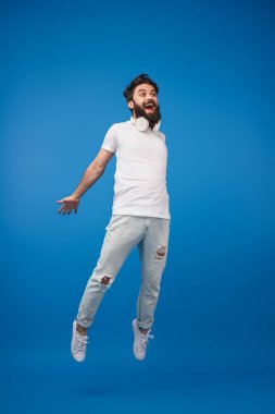 Neşeli genç bir adam havada coşkuyla atlıyor, günlük moda kıyafetleri ve kulaklıklar takıyor, parlak mavi bir stüdyonun arka planına kurulmuş. Neşe, enerji, gençlik gibi kavramlar için mükemmel.