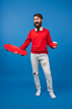 Heyecanlı bir adam elinde kaykay ve kahve tutarken, çarpıcı bir mavi arka plan, enerji iletimi, gençlik ve dinamik bir şehir yaşam tarzına yakalanmış..