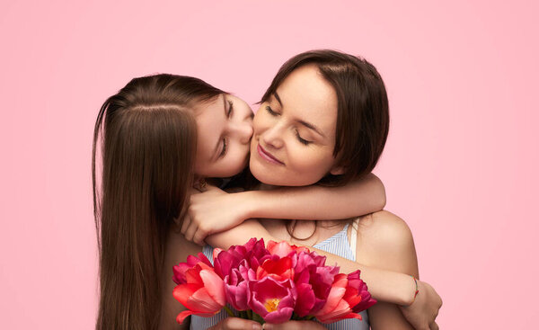 Маленькая девочка целует весёлую женщину в щеку, преподносит букет тюльпанов на весенний праздник на розовом фоне
