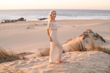 Gün batımında kumlu bir sahilde narin, dantelli bir elbise giymiş bir kadının huzur ve zarafeti yakaladığı huzurlu bir görüntü. Okyanus dalgaları yumuşak, resimsi bir zemin oluşturur..