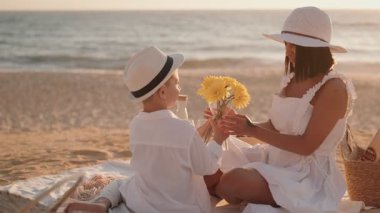 Bir anne ve oğlu güneşli bir günde keyifli bir plaj pikniğinin tadını çıkarırlar. Genç çocuk annesine bir buket sarı çiçekle sürpriz yaparak sevgi ve beraberlik dolu bir an yaratır..