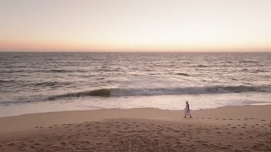 Gün batımında kumsalda tek başına yürüyen bir kadının hava görüntüsü. Derin düşüncelere dalmış ve dalgalar arkasına çarpıyor..