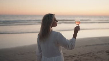 Beyaz elbiseli sarışın bir kadın, okyanusun üzerinde güneş batarken kumsalda bir bardak gül şarabı içiyor. Bu video huzur, sükunet ve gevşemeyi çağrıştırıyor..