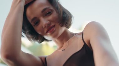 Çekici, şık, genç bir kadın kameraya bakarken gülümseyip saçıyla oynuyor. Aşağıdan doğal ışıklandırmayla çekilmiş..