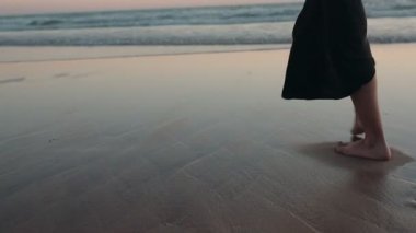 Siyah elbiseli bir kadın gün batımında sahilde yürüyor, dalgalar yavaşça kıyıya vuruyor. Seyahat, tatil ya da farkındalık videoları için mükemmel.