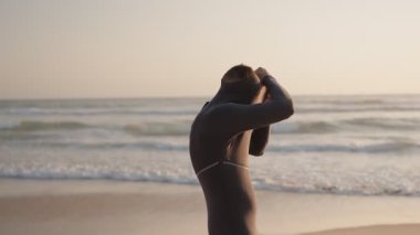 Bir sörfçü gün doğumunda sahilde dalgıç kıyafetini giyer ve sabah sörf seansına hazırlanır. Fermuarını çeker ve elbisesini ayarlar, dalgaları yakalamak için hazırdır..