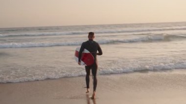 Dalgıç kıyafetli bir sörfçü gün batımında sörf tahtasıyla okyanus dalgalarına doğru koşar. Dalgaları yakalamak için sabırsızlanıyor..