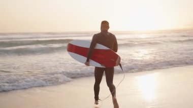 Dalgıç kıyafetli bir sörfçü gün batımında sörf tahtasıyla okyanusa doğru koşar. Sörf yapmaya hazır..
