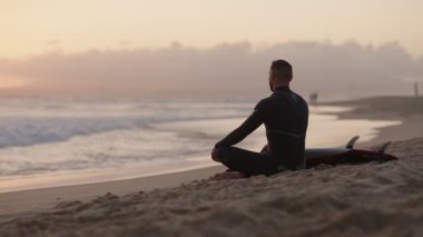 Dalgıç kıyafetli bir sörfçü gün batımında sahilde meditasyon yapar. Sörf tahtasıyla yan yana oturur ve huzurlu anın tadını çıkarır..