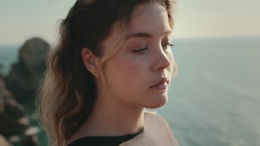 Yansıtıcı genç bir kadın, arka planda deniz olan kameraya bakar. Düşünce ve sükunet hissi uyandırır..