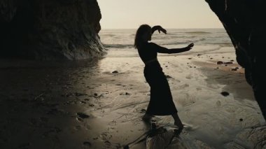 Zarif bir bayan dansçı, gün batımında sakin bir plajda, okyanus kıyısındaki özgürlüğü ve zarafeti simgeleyen siluet dansı yapar..