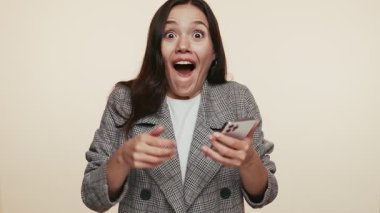 Ekose ceketli neşeli genç bir kadın, elinde muzaffer bir jestle elinde akıllı bir telefonla mutluluğu haykırıyor..