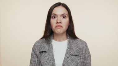 20 'li yaşlarda genç bir esmer, bej bir arka planda damalı ceket giymiş, komik kızgın bir ifade sergiliyor..