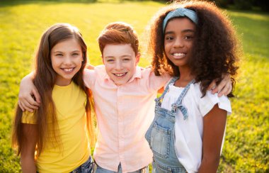 Üç neşeli çocuk birlikte poz veriyor. Parktaki sıcak güneşin altında arkadaşlıklarını ve mutluluklarını gösteriyorlar..