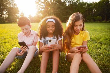 Parkta çimenlerde oturan üç çocuk altın ışıkta akıllı telefonlarının keyfini çıkarıyorlar. Açık hava ortamında teknolojiyi kullanan bağlantılar, oyunlar ve gençleri gösteren samimi bir sahne..