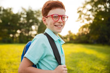 Kırmızı gözlüklü ve sırt çantalı neşeli genç bir çocuk yeşil park ortamında güneşli bir günün tadını çıkarıyor. Canlı renkler ve neşeli ifade, mutluluğu ve açık havaya olan sevgiyi ifade eder..