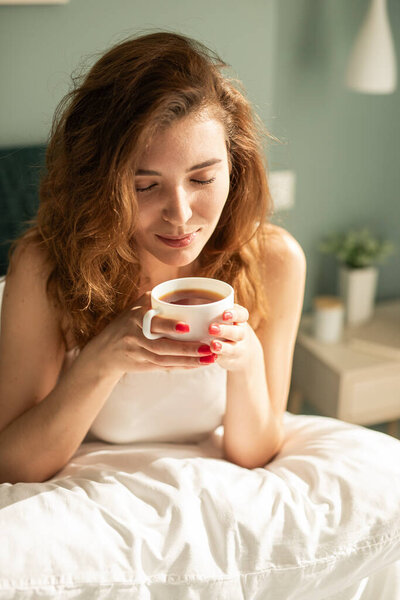 A serene moment featuring a woman joyfully holding a cup of tea in a cozy bedroom setting, emphasizing relaxation, tranquility, and warmth, perfect for morning rituals or peaceful indoor scenarios.