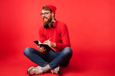 Gündelik giysiler içinde neşeli bir genç adam ve bir deftere yazan gözlükler. Kırmızı bir kazak giyiyor ve çapraz bacaklı oturuyor. Kırmızı arka plana karşı neşeli ve rahat bir atmosfer yaratıyor..