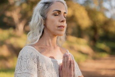 Dışarıda sakin bir ortamda meditasyon ve farkındalık yapan yaşlı bir kadın. Sıcak güneş ışığı ve huzurlu çevre rahatlamayı ve odaklanmayı arttırır. Huzur ve sükunet çok açık.