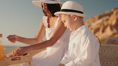 Bir anne ve küçük oğlu güneşli bir plajda aile tatilinin tadını çıkarıyorlar. Beyazlar içinde kumda bir şeyler yiyorlar..