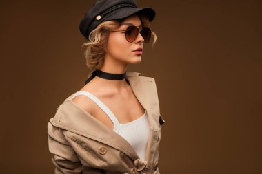 Güneş gözlüğü, şapkası ve bej renkli trençkotlu şık bir kadın. Modern moda eğilimlerini ve kendine güveni sunan şık bir görünüş, çağdaş zarafeti sergilemek için mükemmel.