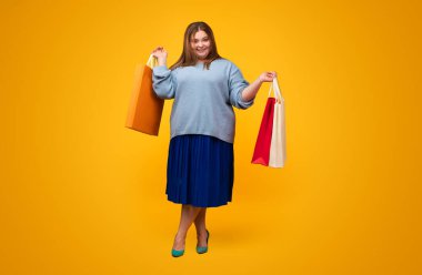 Sarı arka planda duran alışveriş torbalarını tutan tombul genç bir kadın vücudu.