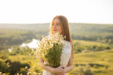 Açık havada elinde bir buket papatya çiçeği tutan, gün ışığının tadını çıkaran ve doğal, huzurlu kırsal manzaralarla çevrili genç bir kadının yer aldığı sakin bir sahne..