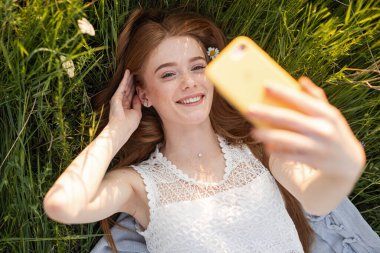 Neşeli bir kadın güneş ışığı altında yeşil çimlere uzanıyor, gülümsüyor ve özçekim anını yakalıyor. Canlı ve taze açık hava havası mutluluğu, eğlenceyi, insan bağlantısını ve güzelliği vurgular.