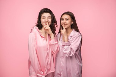 Pembe saten pijama giyen iki neşeli genç kadın, neşeli bir şşşş hareketi yapıyorlar, pembe bir arkaplanda durup gülümsüyorlar ve keyifli ve rahat bir anda eğleniyorlar..