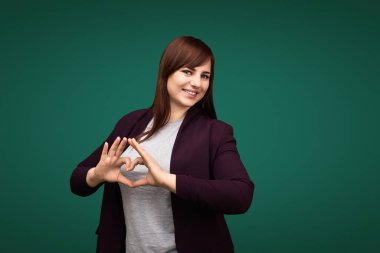 Kendinden emin, iri yarı bir kadın elleriyle kalp şekli oluşturuyor, pozitifliği ve özeni sembolize ediyor. Minimalist yeşil bir arka planda yakalanmış ve sıcak ve dostane bir ifadeye sahip..