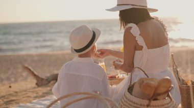 Anne ve oğlu plajda pikniğin tadını çıkarıyor, yemeklerini paylaşıyor ve yaz günbatımının tadını çıkarıyorlar..