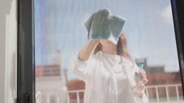 Neşeli genç bir kadın ev işleriyle meşgul, pencereyi paçavra ve temizlik spreyiyle siliyor, parlak bir ev ortamında temizlik konseptini sergiliyor..