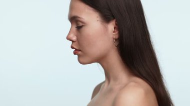 Açık tenli, zarif bir gülüşü olan yakın plan genç bir kadın, açık arkaplanda kameraya doğru dönüyor. İfadesi kendine güven ve doğal güzellik yayar..