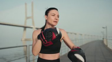 Boks eldivenli kararlı bir kadın antrenörle antrenman yapıyor. Tahta kaldırımda ped kullanıyor. Spor ve adanmışlığı gösteriyor..